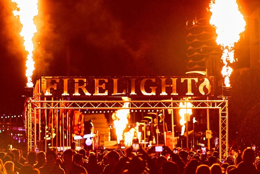 Large lettering saying FIRELIGHT above a stage at the Firelight Festival with two large flames shooting into the air at either end. 