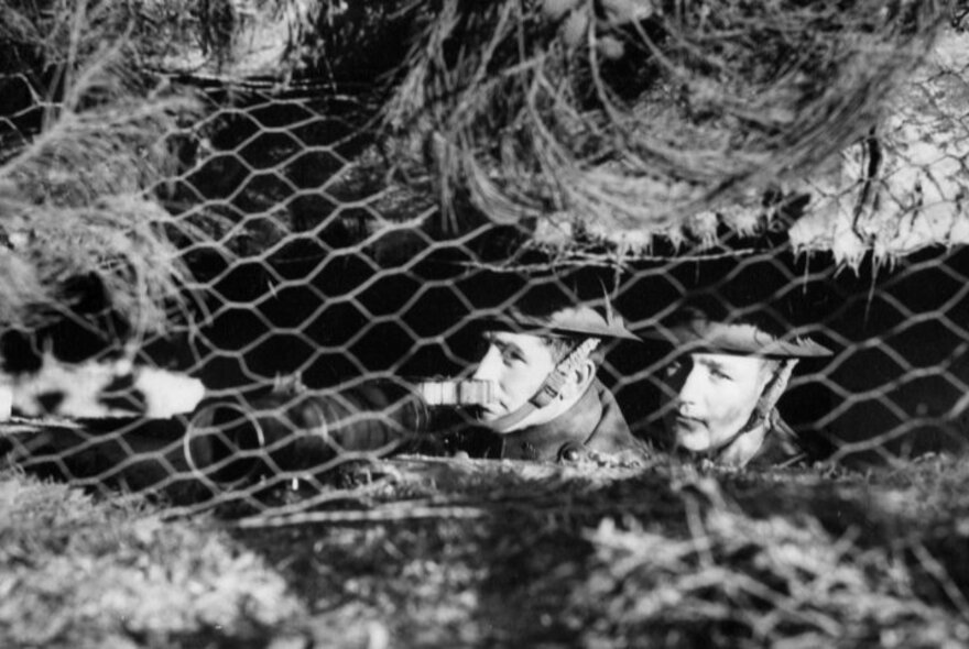 A black and white image of two WWII soldiers in a trench covered with chicken wire.