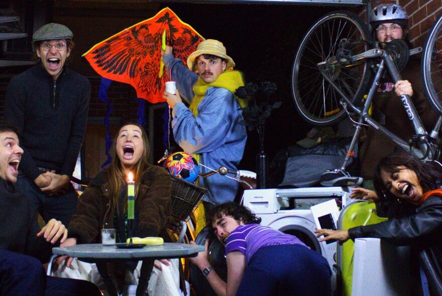 Collection of young performers on a theatre stage all in different poses, one screaming and looking upwards, one holding an umbrella, one carrying a bicycle, one reaching into a front loading washing machine.