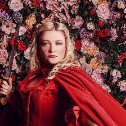 Cast member of opera production of Don Giovanni, a woman with blonde hair wearing a red dress and cape, holding a small knife stands in front of a wall of dense, colourful flowers.