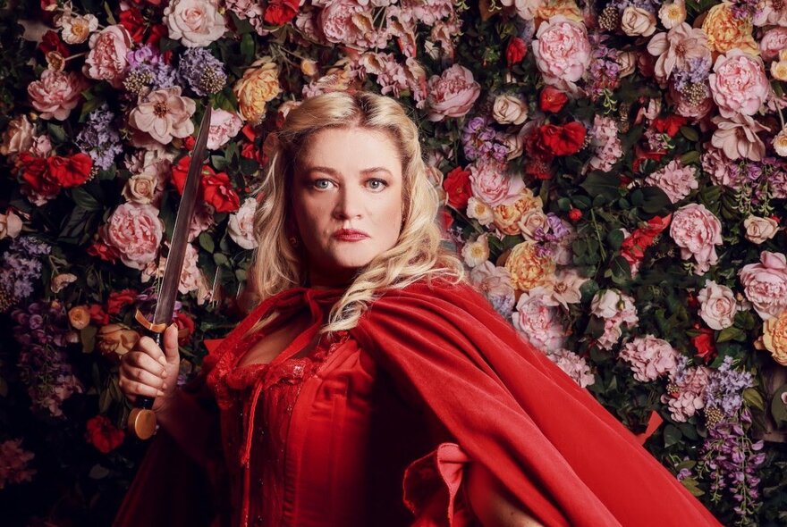 Cast member of opera production of Don Giovanni, a woman with blonde hair wearing a red dress and cape, holding a small knife stands in front of a wall of dense, colourful flowers.