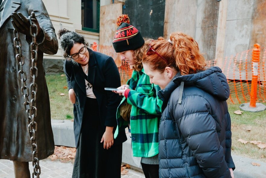 A tour guide talking near a bronze statue, with two tour group participants listening and taking photos. 