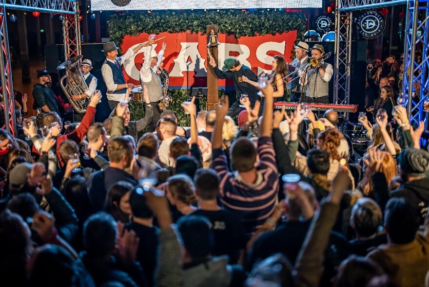 Crowds cheering people on stage with glasses of beer and a brass band.