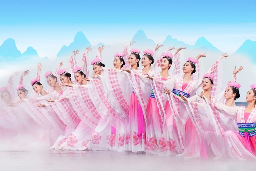 Traditional Chinese dancers in long pink dresses all performing in formation with a set depicting mountains. 