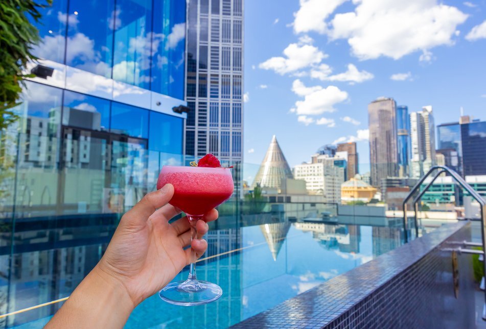 A person is holding a cocktail at a rooftop pool