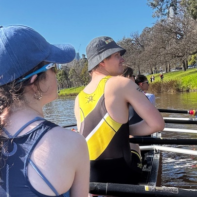 People rowing in a boat, seen from behind, with the river and trees in the background.