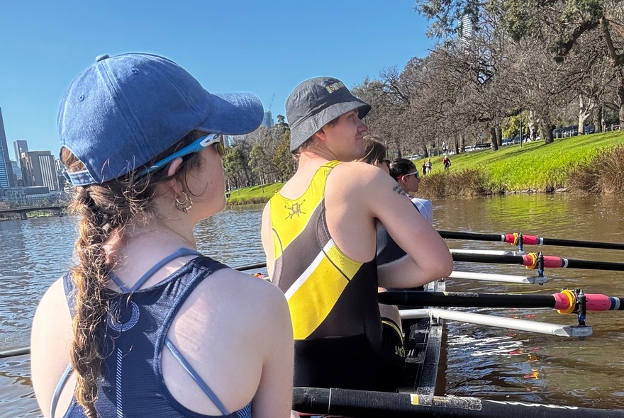 People rowing in a boat, seen from behind, with the river and trees in the background.