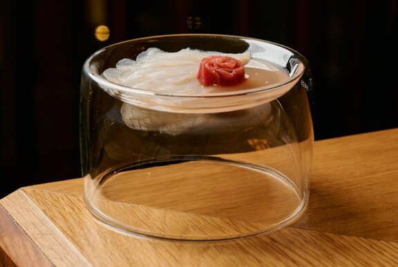 An inverted glass dish on a wooden table, with wafer-thin slices of fugu pufferfish on the top.
