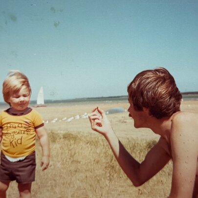 A young child wearing a yellow t-shirt and dark shorts standing near a seated, shirtless individual on a sandy beach or coastal area with a clear blue sky.