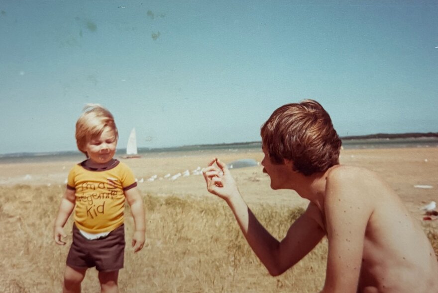 A young child wearing a yellow t-shirt and dark shorts standing near a seated, shirtless individual on a sandy beach or coastal area with a clear blue sky.