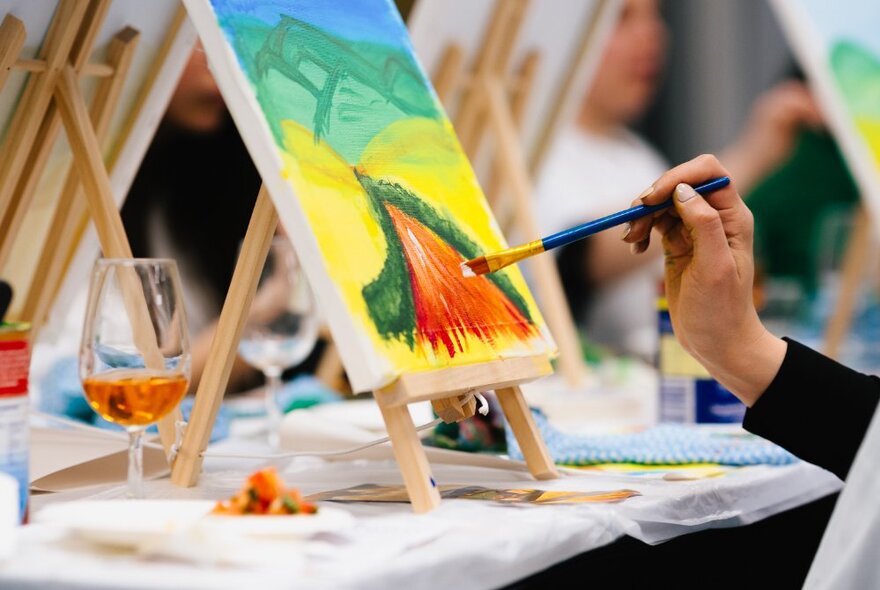 A small canvas with a colourful painted landscape on a tabletop easel; a hand holding a paint brush to the canvas.