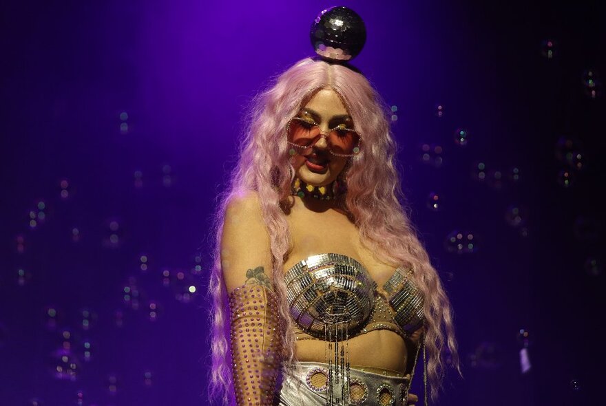 A burlesque performer wearing a silver bustiere, pink sunglasses and a long pink wig, on a darkened stage.