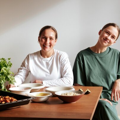 Two young women sit smiling at a wooden dining table set with bowls of food, a baking dish, and a green potted plant.