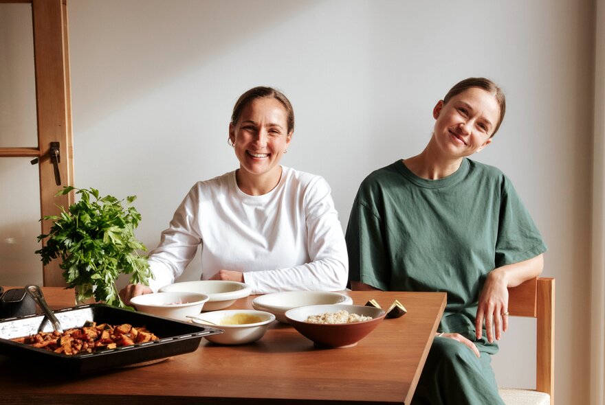 Two young women sit smiling at a wooden dining table set with bowls of food, a baking dish, and a green potted plant.