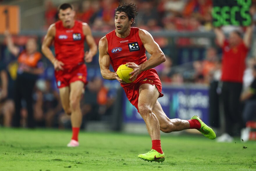 Two Gold Coast SUNS AFL players on the field, one of them running with the ball, blurred footy fans in the stadium in the background.
