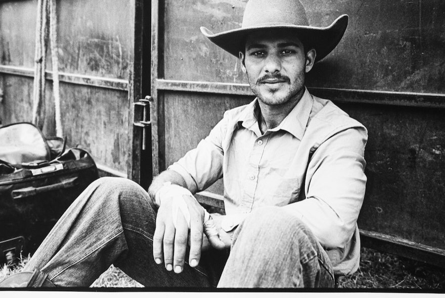 Art photo of a man wearing a hat seated on the ground next to a metal gate.