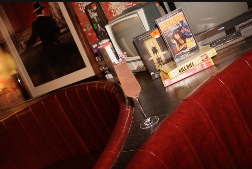 Angled shot of a champagne glass resting on a bench between two leather seating booths, with vintage video tapes displayed behind it.