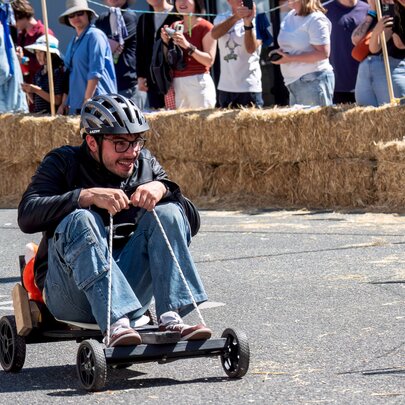 A man wearing a helmet and riding a small billy cart on the street past a crowd behind a hay bale barricade.