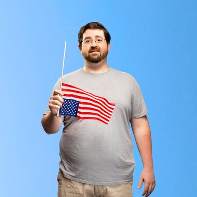 Comedian Richard Mealey wearing a grey t-shirt and holding a small flag of the USA upside down in his hand, posed against a sky blue background.