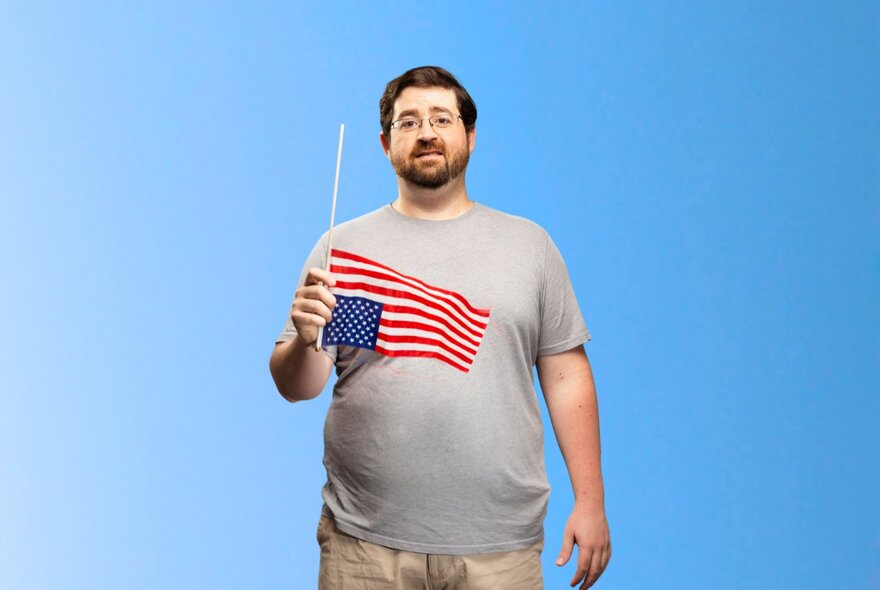 Comedian Richard Mealey wearing a grey t-shirt and holding a small flag of the USA upside down in his hand, posed against a sky blue background.