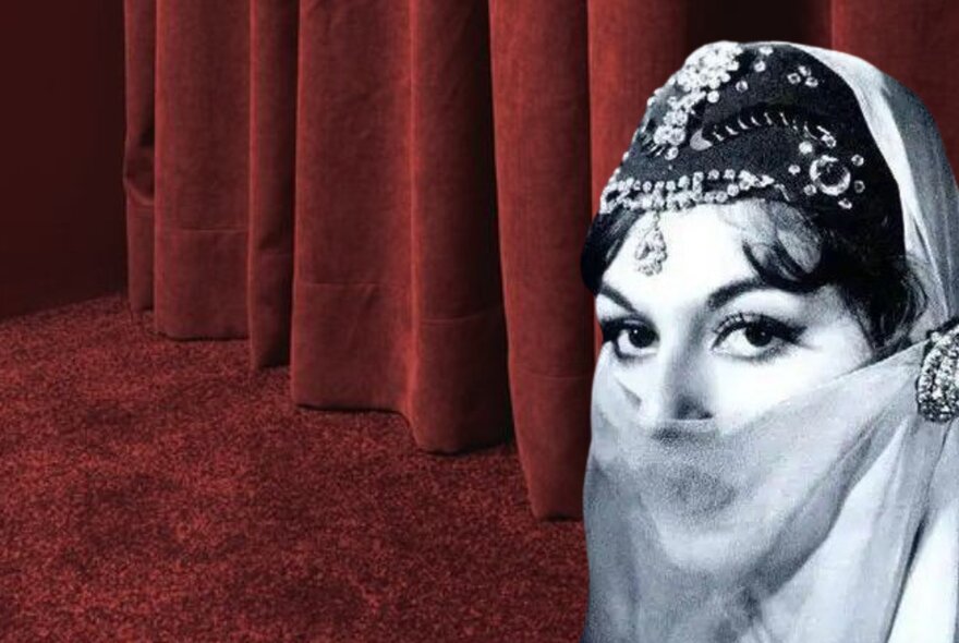 Black and white photo of a woman's face, wearing a beaded headpiece and a half veil over her mouth, with a red velvet curtain and red carpet in the background.