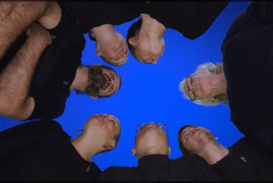 A huddle of people wearing black, seen from below in the centre of the huddle, with deep blue sky above.