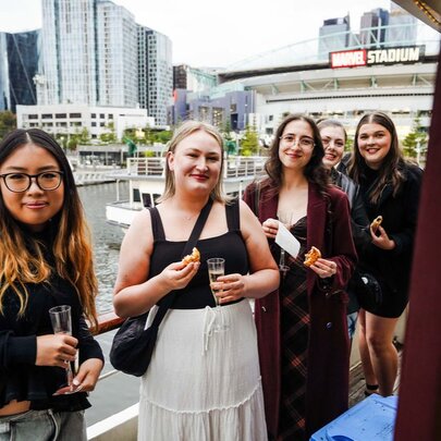 Women holding glasses of bubbles while standing on the deck of a boat moored in Docklands.