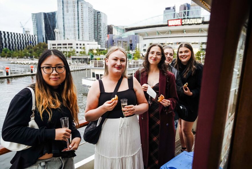 Women holding glasses of bubbles while standing on the deck of a boat moored in Docklands.