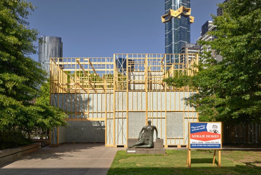 The NGV Architecture Commission showing a timber external house frame on site in the gardens, with trees and blue sky visible, a advertising placard on the lawn in front of the structure.