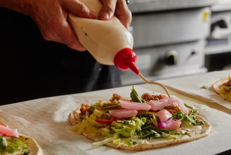 Two hands squeezing a sauce bottle, spreading tahini sauce on a Lebanese wrap.