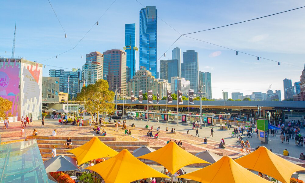 A view of Fed Square on a sunny day with yellow umbrellas in the foreground and the city skyline in the background.