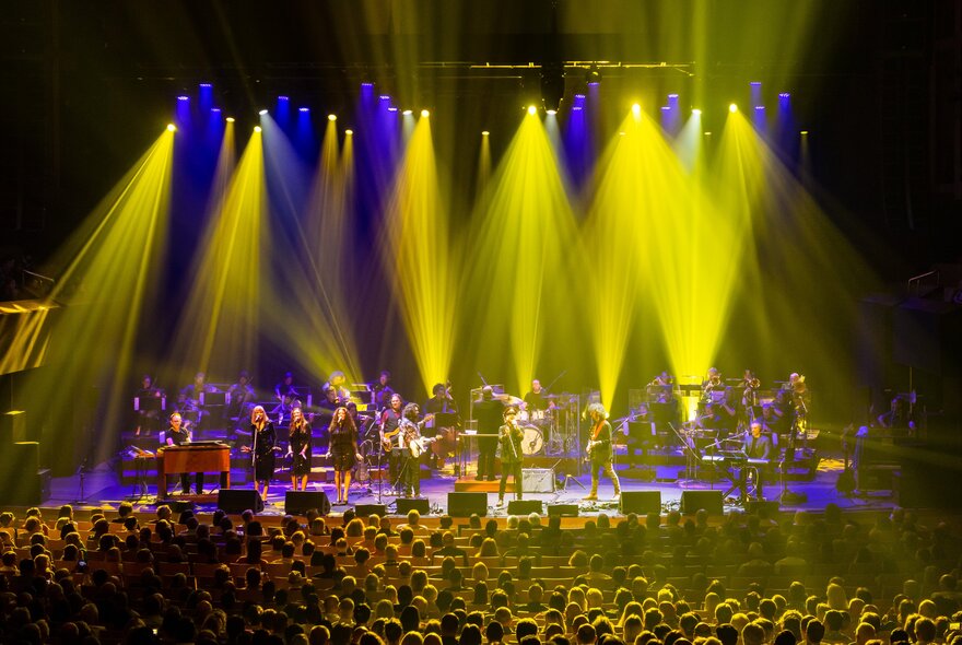 Large rock ensemble performing on stage under purple and yellow lights with seated audience.