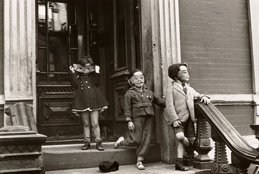 A black and white photograph of three young children on the steps of a building, each wearing a paper mask, circa 1920.