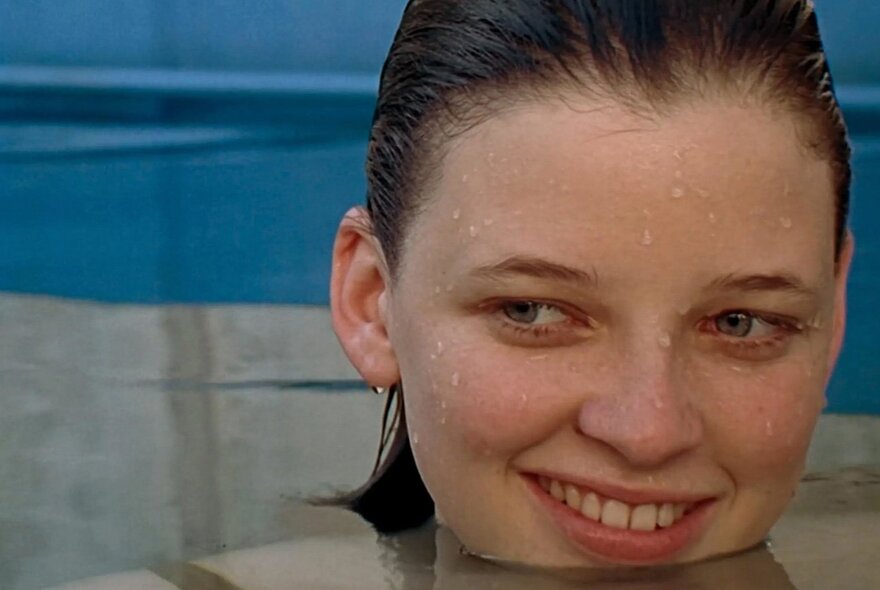 A still from a film, of a young person's smiling head above the water's surface in a swimming pool.