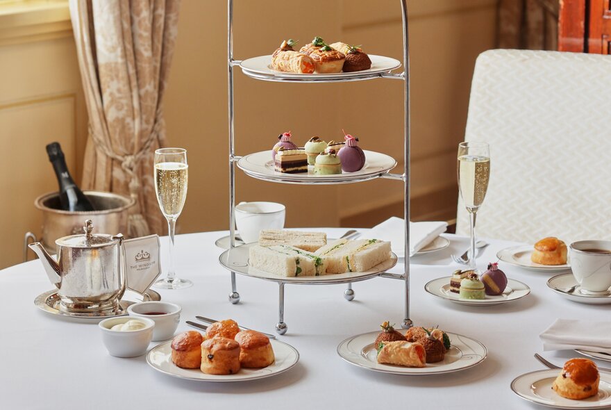 A white table with a three-tiered high tea stand surrounded by small plates of snacks, mugs and champagne.