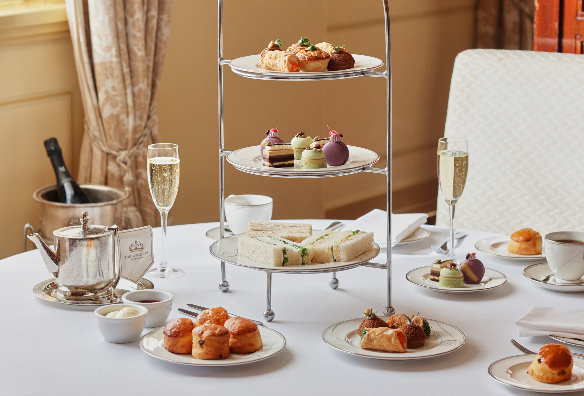 A white table with a three-tiered high tea stand surrounded by small plates of snacks, mugs and champagne.