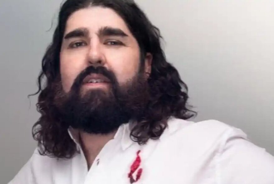 Man with black beard and shoulder-length hair, wearing a white shirt with red detail.