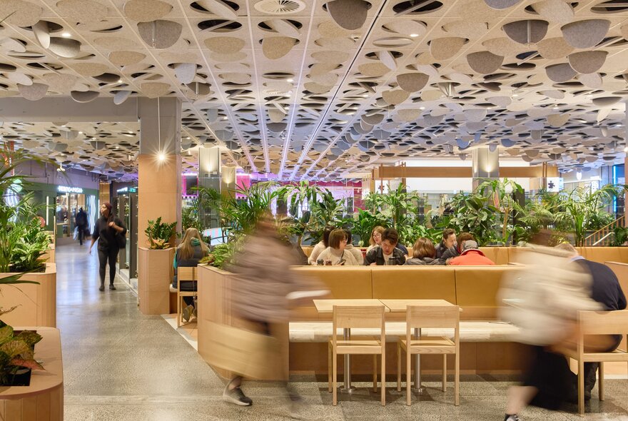 Food court with wooden seats and tables, with blurred figures.