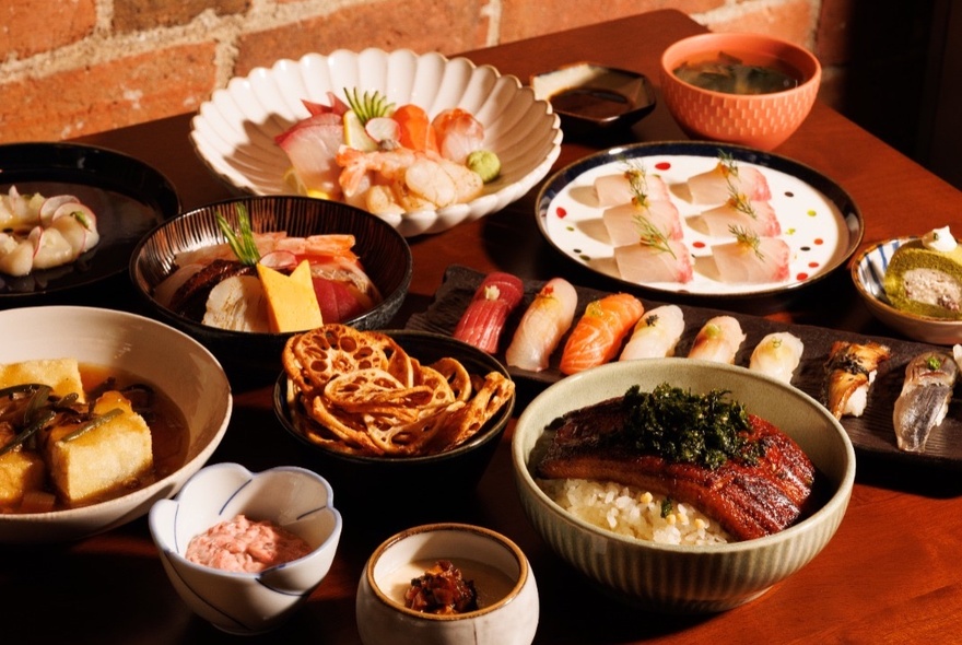Array of Japanese food in different sized ceramic bowls and plates, on a small wooden dining table.