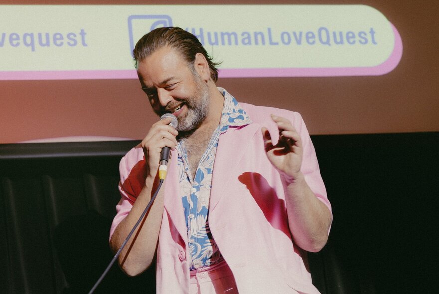 Bearded man holding a microphone, wearing a pink jacket and patterned blue shirt.