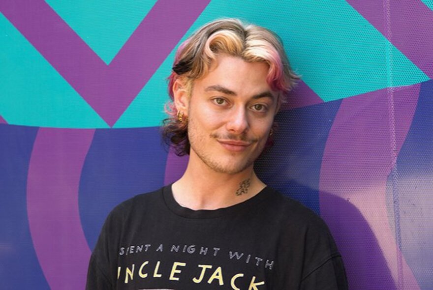 A man with curled, streaked hair and a small moustache, smiling against a coloured background. 
