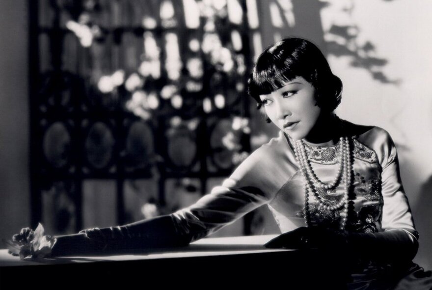 An old black and white image of a glamorous Anna May Wong with a 1920s bob and many strings of pearls around her neck. 