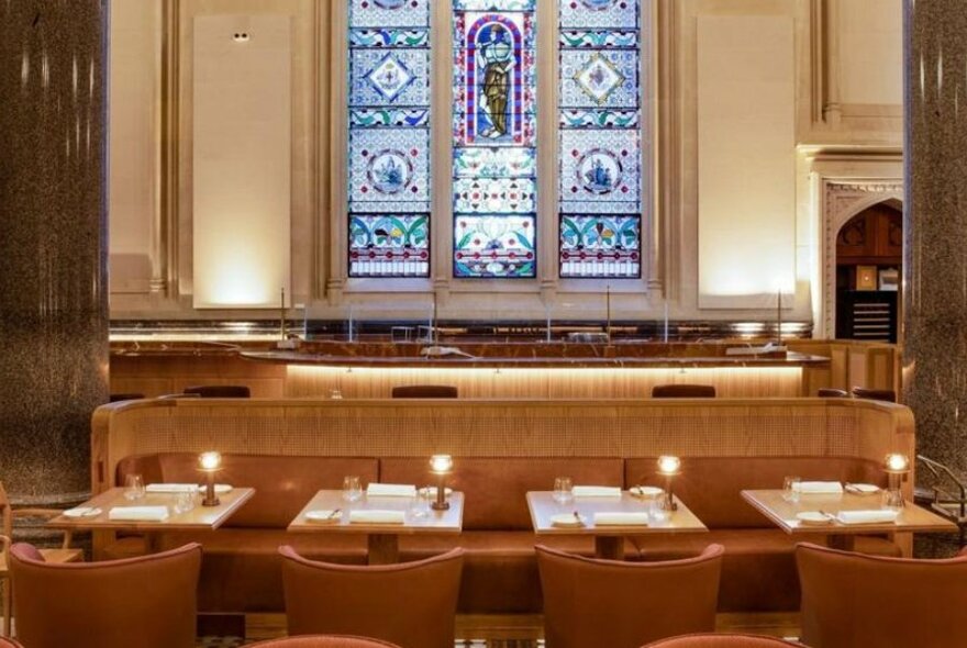 Interior view of La Reine restaurant showing banquette seating, small tables set for two and club chairs, large marble columns and a rear wall with a stained glass feature window.