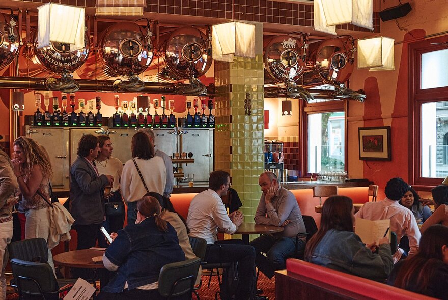 People standing at a bar and seated at tables in a drinking venue.