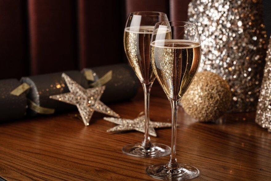 Two champagne flute glasses filled with sparkling wine, with festive gold and silver Christmas decorations in the background.