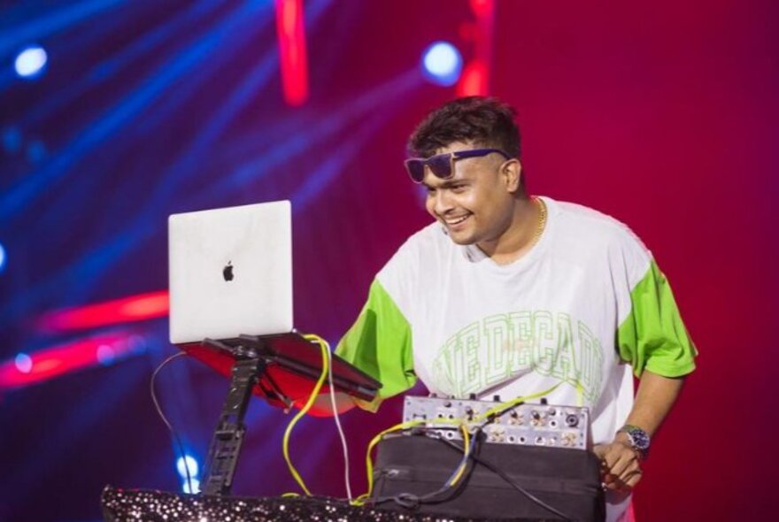 A smiling DJ performing in front of a MacBook with sunglasses on his forehead.