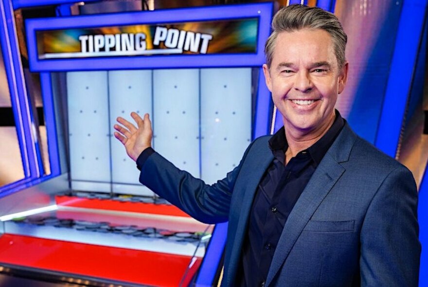 Todd Woodbridge smiling and waving his arm towards a Tipping Point arcade game display in the background.