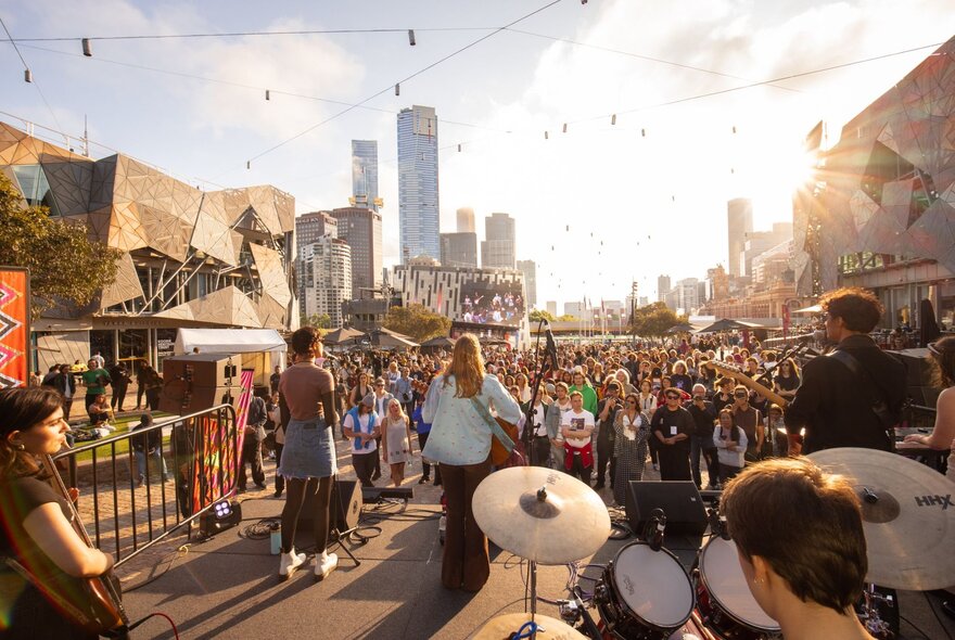 A young band with multiple musicians on stage at Fed Square, seen from behind and looking into the afternoon sun. 