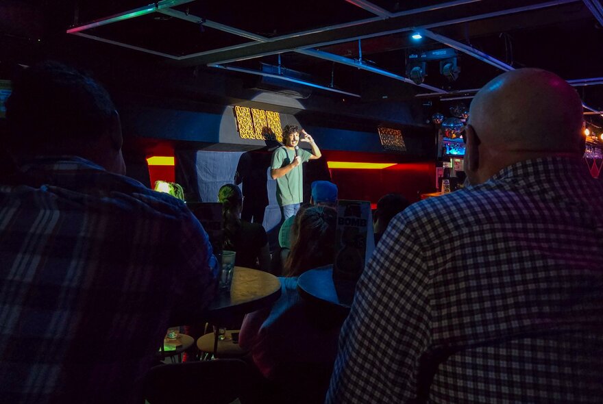 A person on a stage, speaking to a seated live audience at a venue hosting stand-up comedy.