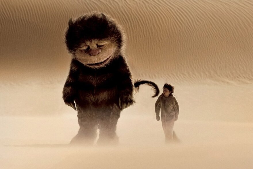 A still from the movie Where the Wild Things Are, showing a giant furry monster with a melancholy looking face, walking along a sand dune with a young boy.